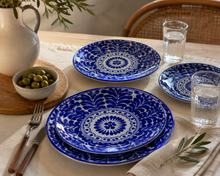 Load image into Gallery viewer, Blue and white ceramic tableware set on a dining table, including two oval plates with intricate floral mandala patterns, a smaller matching plate, clear drinking glasses, silverware, a bowl of olives, and a white pitcher with olive branches.
